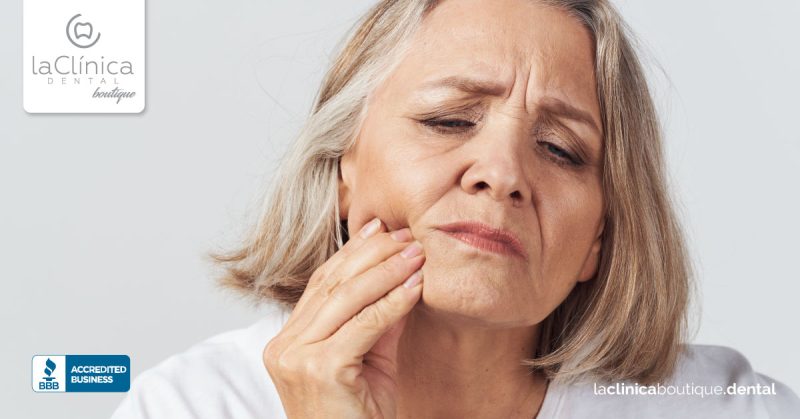Mujer mayor con dolor dental, toc&aacute;ndose la mejilla, representando la importancia de la salud dental en La Cl&iacute;nica Dental Boutique.