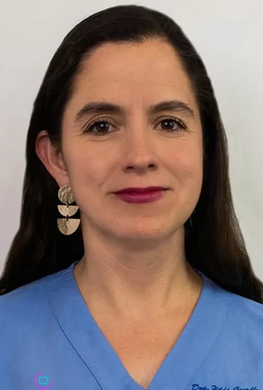 Mujer con cabello largo y aretes, vistiendo una bata azul, retrato de la Dra. Hilda G., especialista en cirugía maxilofacial en La Clínica Dental Lindavista.