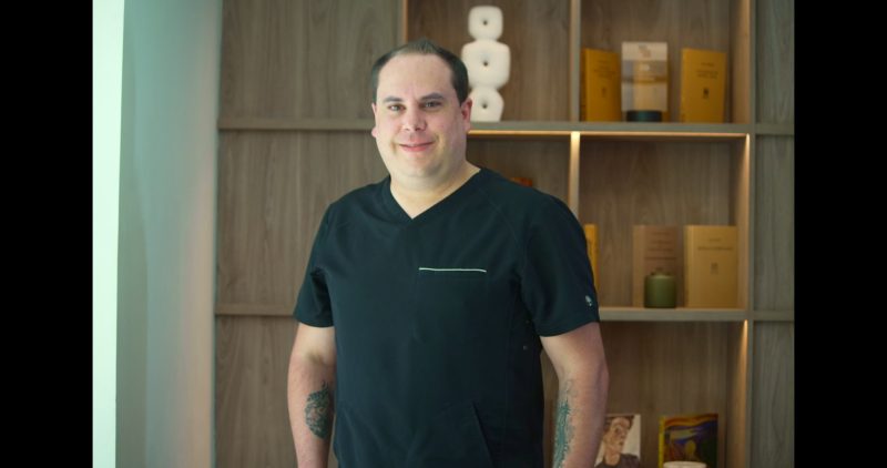 Dentista en uniforme negro sonriendo, con fondo de estantería que muestra productos y documentos, representando el equipo profesional de La Clínica Dental Lindavista.