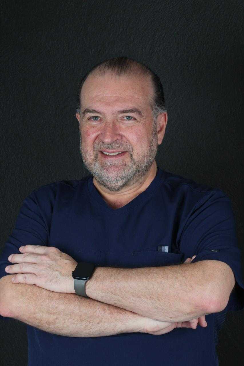 Dr. Sergio M. smiling with crossed arms, wearing a navy blue dental uniform, against a dark background, representing expertise in periodontics and implantology at La Clinica Dental Lindavista.